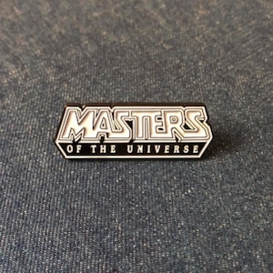 May include: A rectangular pin featuring the words "MASTERS OF THE UNIVERSE" in white with a black outline. The pin is set against a textured, dark blue background. A collectible item.