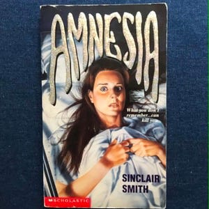 May include: A paperback book titled Amnesia by Sinclair Smith with a Scholastic logo. The cover features a person with wide eyes clutching a light blue blanket. The title is written in large, textured silver letters, and the text reads: What you don't remember...can kill you.