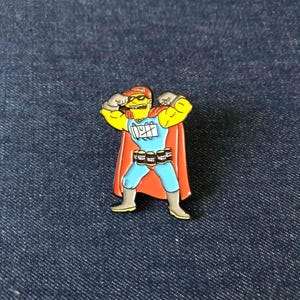May include: A colorful enamel pin featuring a cartoon character in a superhero costume. The character is yellow with a red hat and cape, blue suit, and holding up his arms. The pin has the word "Duff" on the chest and beer cans on the belt.