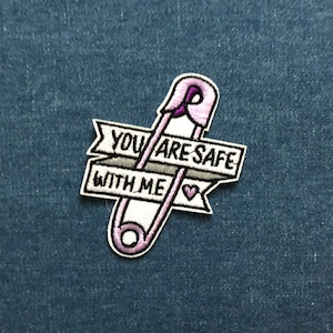 May include: Embroidered patch with a pink safety pin design and the text "YOU ARE SAFE WITH ME" in black. The patch has a white border and a small heart symbol. The background is a textured blue fabric.