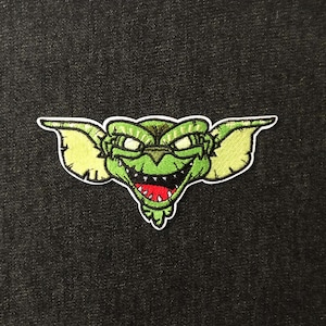 May include: Embroidered patch featuring a green creature with large ears, a wide grin revealing red teeth, and white eyes. The patch has a white border and is set against a dark background. This patch is perfect for adding to jackets, bags, or other fabric items.