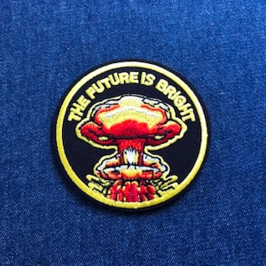 May include: A circular patch with the text "THE FUTURE IS BRIGHT" encircling a red and yellow mushroom cloud design on a black background. The patch is on a blue denim surface.