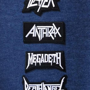 May include: Four embroidered patches with heavy metal band logos: Slayer, Anthrax, Megadeth, and Death Angel. Each patch has a black background with white lettering and a white border. The patches are displayed on a blue denim background.