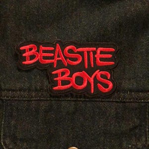 May include: Dark denim jacket featuring a red and black embroidered "BEASTIE BOYS" patch. The patch is sewn onto the pocket area. A single, round, bronze-coloured button is visible on the jacket.