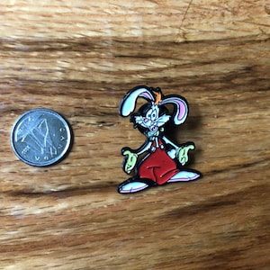 May include: A cartoon rabbit enamel pin with white, pink, and black details, wearing red overalls. A coin is next to the pin for size comparison. The pin is on a wooden surface.