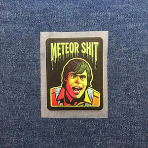 May include: A rectangular embroidered patch featuring a graphic of a person with a surprised expression. The text METEOR SHIT appears in a dripping lime green font above the person, who wears a collared shirt and vest against a black background.