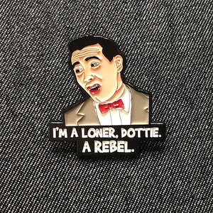 May include: A black metal enamel pin featuring a cartoon illustration of a man with dark hair, wearing a tan suit jacket, white shirt, and red bow tie. Below the figure, black text on a rectangular base reads: I'M A LONER, DOTTIE. A REBEL.