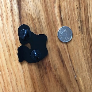 May include: Black enamel pin with two pin backs, next to a silver coin. The pin has a unique shape. The coin is a quarter. The pin and coin are on a wooden surface.