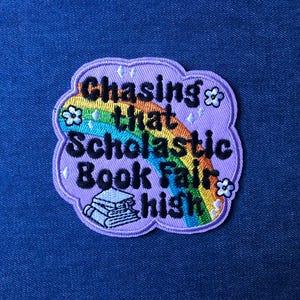 May include: A lilac embroidered patch with the text "Chasing that Scholastic Book Fair high" in black, with a rainbow and flower accents. A stack of books is at the bottom. The patch is on a blue denim background.