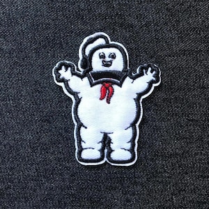 May include: A white embroidered patch featuring the Stay Puft Marshmallow Man from Ghostbusters. The character has a black sailor hat and collar, a red neckerchief, and a smiling face. The patch is outlined in black.