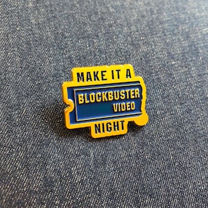 May include: A rectangular enamel pin featuring a yellow border and a blue centre. The text reads "MAKE IT A BLOCKBUSTER VIDEO NIGHT" in yellow and blue lettering. The design is shaped like a ticket stub with a notched edge on the left side.