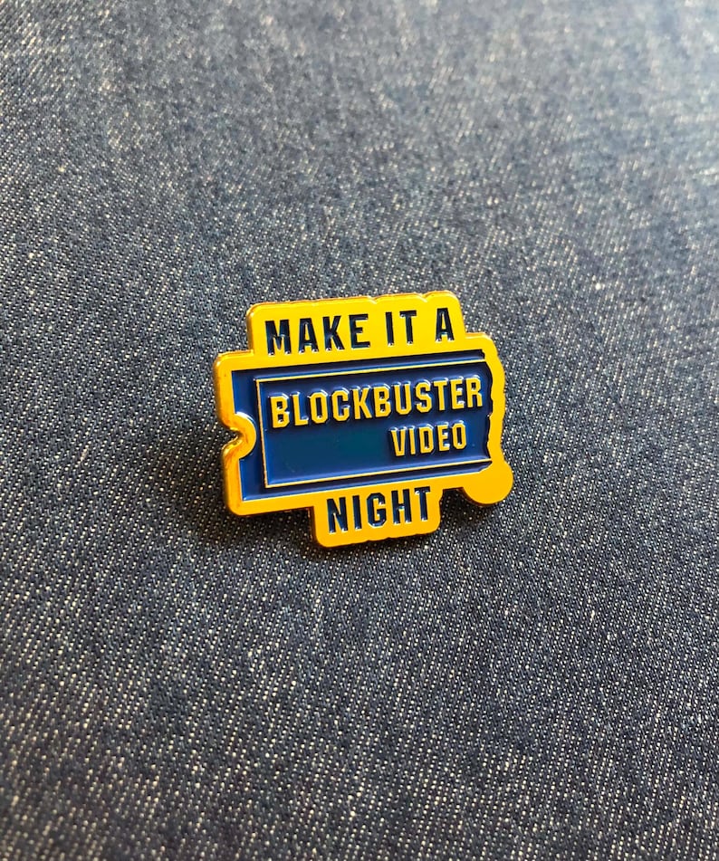 May include: A rectangular enamel pin featuring a yellow border and a blue center. The text reads "MAKE IT A BLOCKBUSTER VIDEO NIGHT" in yellow and blue lettering. The design is shaped like a ticket stub with a notched edge on the left side.