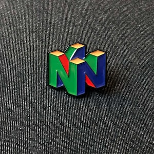 Peut inclure: Une épingle colorée en émail représentant le logo Nintendo 64. L'épingle présente un design en trois dimensions avec les lettres "N" en vert et bleu, et un accent rouge. Le dessus des lettres est doré.