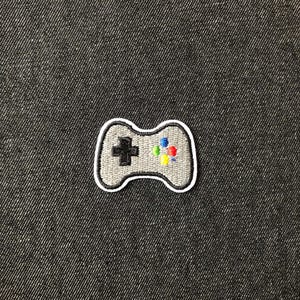 May include: A grey embroidered patch of a video game controller. The controller features a black cross-shaped directional pad and colourful buttons. The patch is outlined in white and is on a dark grey textured background.