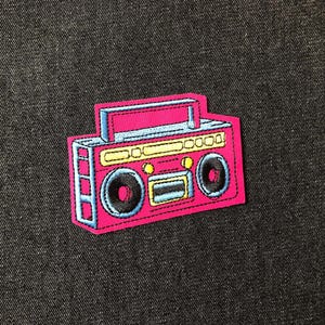 May include: A pink embroidered patch in the shape of a retro boombox. The patch features a blue handle, speakers, and accents, with yellow and black details. The patch is set against a dark denim background.