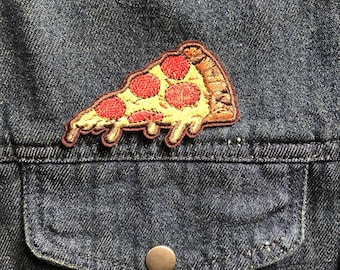 May include: Embroidered pizza slice patch with red pepperoni, yellow cheese, and a brown crust. The patch is sewn onto a dark blue denim jacket, positioned above a pocket with a silver button.
