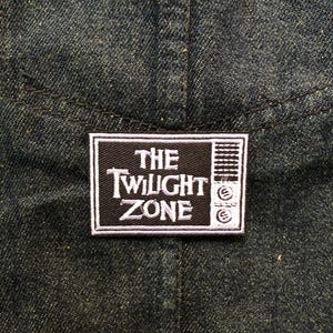 May include: A dark denim fabric with a rectangular patch featuring the text "THE TWILIGHT ZONE" in white lettering. The patch has a black background and a white border, with a stylized television graphic.