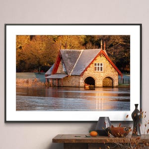 May include: A stone boathouse with a red roof and red trim, reflected in a still body of water. The boathouse has two arched doorways and a single window. The water is a light blue color and the sky is a light blue color. The boathouse is surrounded by trees with fall foliage.