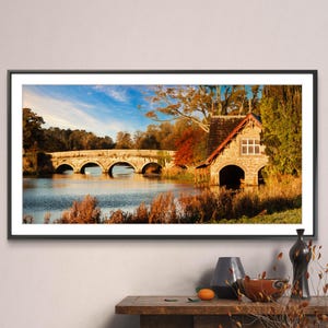 May include: A framed landscape photograph of a stone bridge over a river with a stone boathouse on the right side of the image. The bridge has five arches and is surrounded by trees with fall foliage. The water in the river is calm and reflects the sky and trees. The boathouse has two large arched openings and a red roof. The photograph is in a black frame.