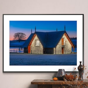 May include: A black framed photograph of a stone building with a slate roof. The building has two windows and a red door. The building is surrounded by a black fence. The building is set against a blue sky with a few clouds. The ground is covered in frost. The photo is displayed on a wooden table with a vase and a bowl.