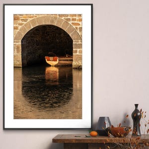 May include: A framed photograph of a small wooden boat docked under a stone archway. The boat is a light brown color with a red stripe. The water is dark blue and reflects the light from the sky. The archway is made of rough stone and is partially in shadow. The photograph is framed in a black frame with a white mat.