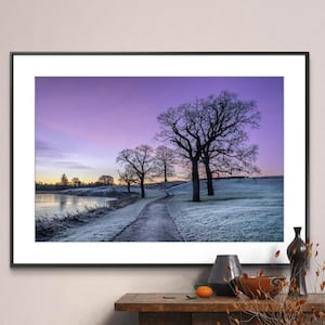 May include: A framed photograph of a purple and pink sunrise over a frozen lake. The lake is surrounded by trees and a path leads away from the lake. The path is covered in frost and the trees are silhouetted against the sky.