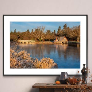 May include: A framed photograph of a lake with a stone bridge and a small stone building with a red roof. The lake is frozen and the water is a deep blue color. The trees surrounding the lake are bare and the sky is a light blue color. The photograph is in a black frame and is hanging on a white wall.