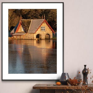 May include: A stone boathouse with a red roof and arched doorways reflected in a still body of water. The boathouse is surrounded by trees and the water is reflecting the golden light of the setting sun.