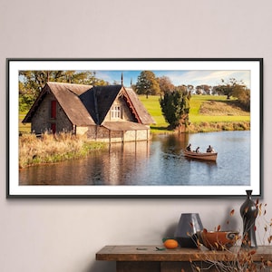 May include: A framed photograph of a stone boathouse with a red roof on a lake. The boathouse is surrounded by green grass and trees. There are three people in a rowboat on the lake.