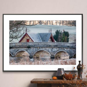 May include: A stone bridge with five arches over a river. A small, red-roofed building with a gray roof is behind the bridge. The scene is set in a wintery landscape with frost on the ground and trees in the background.
