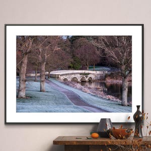 May include: A framed photograph of a white stone bridge over a pond. The bridge has five arches and is surrounded by trees with bare branches. The ground is covered in frost and there is a path leading to the bridge.