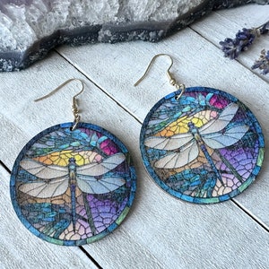 Könnte beinhalten: Runde Holzohrringe mit einem Buntglas-Libellen-Design. Die Ohrringe zeigen eine farbenfrohe Libelle mit blauen, lila, gelben und grünen Akzenten. Die Ohrringe haben silberfarbene Ohrhaken.