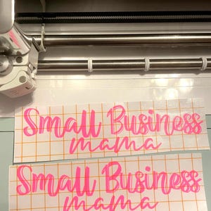以下が含まれることがあります： ピンク色の「Small Business mama」という文字が筆記体で書かれた2枚のビニールデカール。デカールは格子模様の裏地の上にあります。画像は上から撮影されており、カッティングマシンにデカールが貼られている様子が写っています。