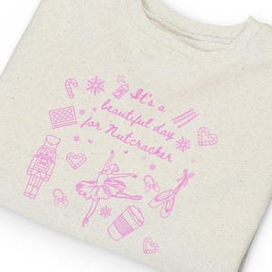 May include: A natural heather crewneck sweatshirt featuring a pink graphic print with the text 'It's a beautiful day for Nutcracker'. The design includes illustrations of a nutcracker, a ballerina, ballet slippers, a coffee cup, and hearts.