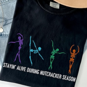 May include: A black short-sleeved t-shirt featuring four colorful skeletons in various ballet poses. The skeletons are purple, blue, green, and orange. Below the skeletons, the white text reads: STAYIN' ALIVE DURING NUTCRACKER SEASON.