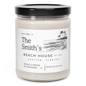 May include: A glass jar candle with a black lid and a white label. The label says "The Smith's Beach House Est. 2024 Jupiter, Florida" and features a black and white illustration of a beach scene with palm trees. The candle is 9 oz and has a burn time of 50-60 hours. The scent is Sea Salt & Orchid.