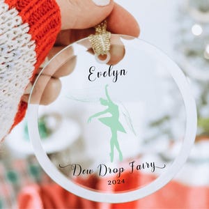 May include: A clear glass ornament with a green silhouette of a ballerina and the text "Evelyn Dew Drop Fairy 2024".