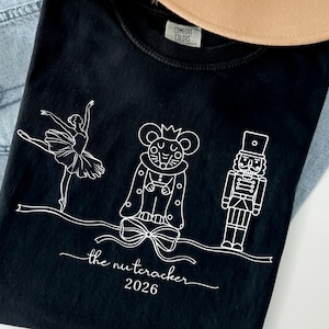 May include: Black t-shirt featuring a white line drawing of a ballerina, a mouse king, and a nutcracker. The text "the nutcracker 2026" is printed below the illustrations. The shirt is folded on top of blue jeans.