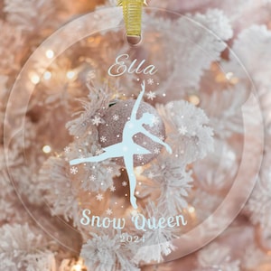 May include: A clear glass ornament with a white silhouette of a ballerina dancing. The ornament has the name "Ella" and the text "Snow Queen 2024" printed on it.