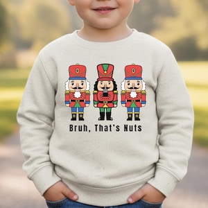 May include: A light beige sweatshirt featuring a graphic of three cartoon nutcrackers in red, green, and blue uniforms. The text "Bruh, That's Nuts" is printed below the nutcrackers. The sweatshirt is worn by a child.