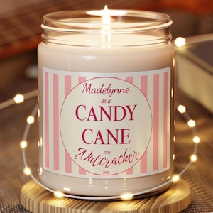 May include: A lit candle in a clear glass jar with a label featuring pink and white vertical stripes. The label text reads Madelynne as a CANDY CANE the Nutcracker 2026. The candle sits on a round wooden base surrounded by a string of small warm lights.