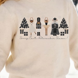 May include: A cream-colored crewneck sweatshirt featuring a graphic print of two Christmas trees, two gift boxes, a ballerina, a nutcracker, a fairy, and a mouse king. The text "Sorry, Cant, Nutcracker Season!" is printed below the figures.