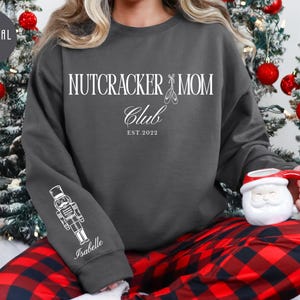 May include: A charcoal gray crewneck sweatshirt featuring white text that reads NUTCRACKER MOM CLUB EST. 2022 with a ballet slipper icon. The right sleeve displays a white line art illustration of a nutcracker and the name Isabelle in script.