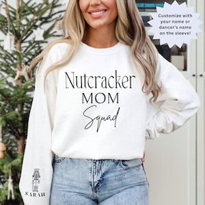 May include: White sweatshirt with the text "Nutcracker MOM Squad" printed on the front. The text is in black. The sweatshirt is being worn by a person. The sleeve has a small image of a nutcracker with the text "SARAH" underneath.