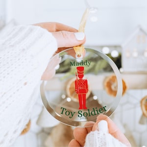 May include: Clear, round Christmas ornament with a red toy soldier design. The ornament has the name "Maddy" and the words "Toy Soldier 2024" in green. A gold ribbon is attached for hanging.