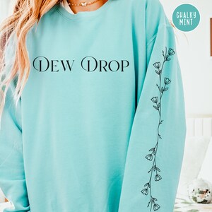 May include: A light blue sweatshirt with the text "DEW DROP" in black. The left sleeve features a black floral vine design. A round sticker in the upper right corner reads "CHALKY MINT".