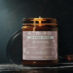 May include: A brown glass candle jar with a white label that says "Serena Paige is giving Snow Queen vibes". The label has a snowflake pattern and the text "The Buttercracker 2024 Christmas Winter Soy Candle 40z" is printed on the bottom.