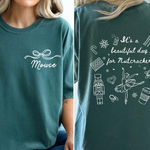 May include: A dark green t-shirt featuring a white graphic on the front chest of a bow and the word Mouse. The back displays white line art of a nutcracker, a ballerina, snowflakes, candy canes, ballet slippers, and the text It's a beautiful day for Nutcracker.