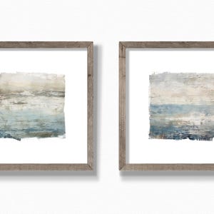 May include: Two framed abstract art prints. Each print features a rectangular composition with layered brushstrokes in shades of blue, beige, and gray. The frames are made of light brown wood, and the artwork is set against a white background.