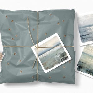 May include: A wrapped gift in teal paper, tied with gold twine and adorned with gold star-shaped embellishments. The package features a small art print of a seascape. Additional seascape art prints are placed nearby, along with a spool of gold twine.